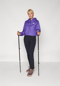 Giacca impermeabile viola con cappuccio, pantaloni neri aderenti e scarpe da trekking marroni. Due bastoni da trekking tenuti in mano. Sfondo pulito e minimalista.