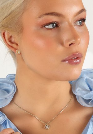 Guldhalsband med en blomsterberlock och diamanter. Modellen bär örhängen med stift och en blå volangblus. Huden har en naturlig lyster.