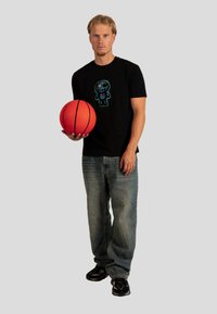 Czarna koszulka z kolorową grafiką kreskówkowego bohatera, w połączeniu z jasnoniebieskimi dżinsami i czarnymi sneakersami, trzymająca żywą czerwoną piłkę koszykową.