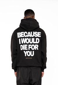 Sudadera con capucha negra que lleva un gran texto blanco en la parte posterior que dice: "PORQUE MORIRÍA POR TI," y un pequeño logotipo dorado en la parte inferior.