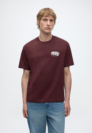 Joven con cabello claro con camiseta gráfica rojo oscuro y vaqueros azules de pie contra un fondo blanco sencillo.