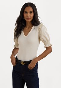 Top de malha canelada em bege claro com decote em V e mangas bufantes. Apresenta delicados detalhes em babados nas bainhas das mangas. Combinado com jeans escuros.