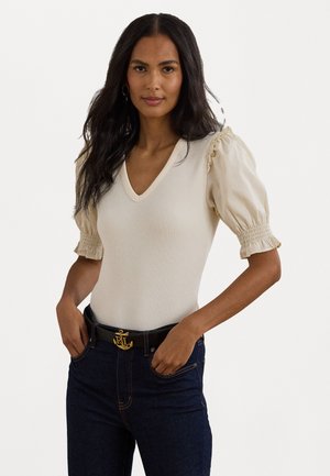 Top a coste beige chiaro con scollo a V e maniche a palloncino. Presenta delicati dettagli a volant sui bordi delle maniche. Abbinato a jeans in denim scuro.