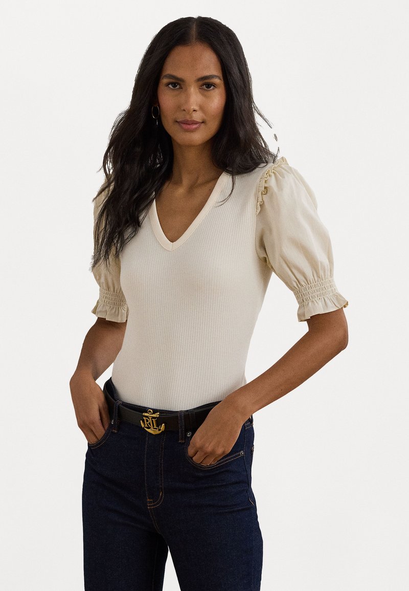 Top de malha canelada em bege claro com decote em V e mangas bufantes. Apresenta delicados detalhes em babados nas bainhas das mangas. Combinado com jeans escuros.