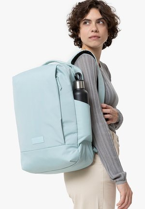 Zaino azzurro chiaro con una texture liscia, chiusura a zip e una tasca laterale che contiene una bottiglia d'acqua nera. Forma rettangolare, spallacci imbottiti.