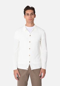 Cardigan bianco lavorato a maglia con colletto, dotato di bottoni in legno sulla parte anteriore, orlo dritto e maniche lunghe. Indossato con pantaloni beige.