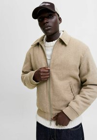 Chaqueta de mezcla de lana beige con cierre de cremallera, cuello puntiagudo y puños acanalados. Combinada con un suéter de punto blanco y jeans de mezclilla oscura.