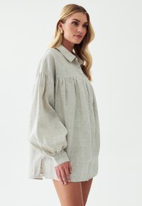 Světle šedá lněná oversized košile s knoflíkovým zapínáním vpředu, nařasenými rameny a nabíranými rukávy. Boční rozparky zlepšují cirkulaci vzduchu a přizpůsobení.