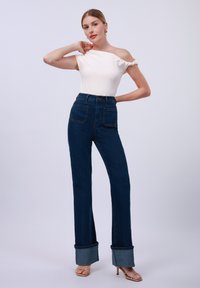 Hochtaillierte, dunkelblaue Jeans aus Denim mit weit ausgestelltem Bein und umgeschlagenem Saum, kombiniert mit einem enganliegenden, elfenbeinfarbenen Oberteil und beigen Riemchen-Heels.