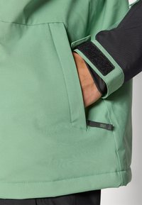 Ανοιχτό πράσινο μπουφάν με λεία υφή, που διαθέτει πλαϊνή τσέπη και μαύρα μανίκια με βέλκρο. Εντυπωσιακές ραφές και μικρή θηλιά ετικέτας.