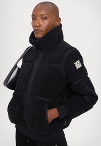 Svart fleecejacka med hög krage, med texturerad tyg, dragkedjestängning och sidofickor. Inkluderar märkeslogotyp på ärmen.