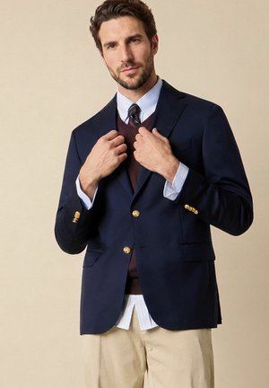Mann richtet marineblauen Blazer mit goldenen Knöpfen, trägt hellblaues Hemd, gestreifte Krawatte, burgunderfarbenen Pullover ohne Ärmel und beige Hose vor einfarbigem Hintergrund.