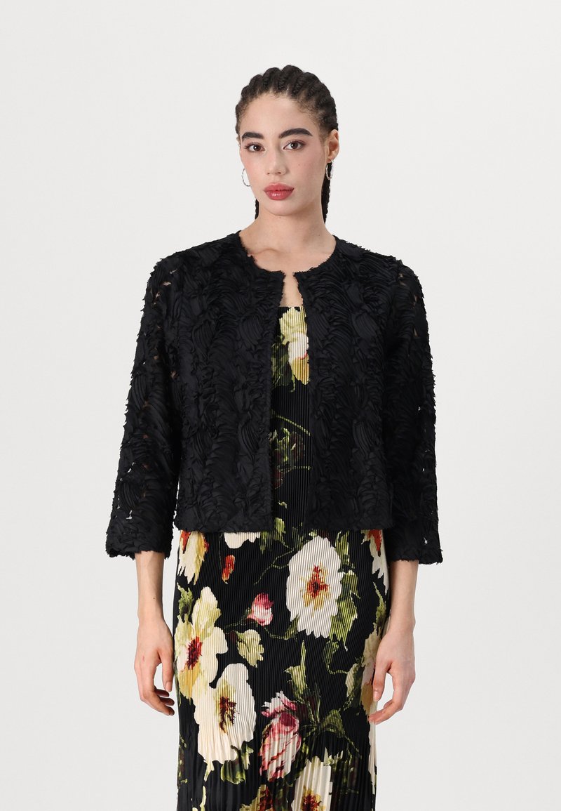 Black embroidered jacket with three-quarter sleeves, textured floral design. Worn over a floral pleated dress featuring white and yellow flowers.