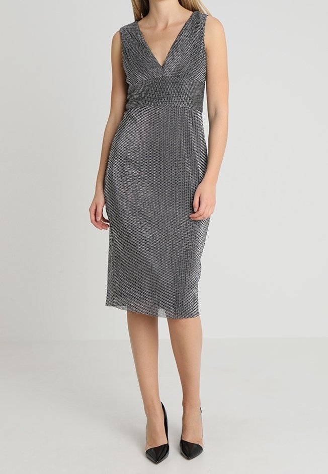 Femme portant une robe noire et blanche à motifs sans manches, longueur genou avec un col en V et des escarpins noirs pointus, debout devant un fond uni.