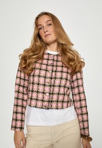 Károvaný tweedový blejzer ružovej a čiernej farby s krátkym strihom, ozdobený guľatými gombíkmi a textúrovaným povrchom. Nosí sa cez bielu košeľu.