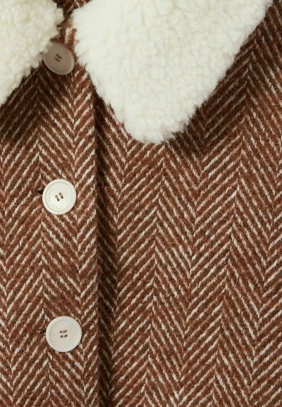 Chaqueta marrón de espiga con cuello de piel sintética blanca, que cuenta con botones redondos y un tejido texturizado y trenzado.