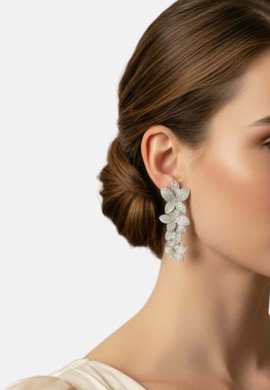 Woman with brown hair in a low bun wearing long, silver floral earrings made of five and four-petal flower clusters on a light background.