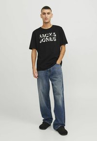 Juodas marškinėliai su balta "JACK & JONES" logotipu, laisvas kirpimas, derinami su šviesiai mėlynomis plačiomis džinsinėmis kelnėmis ir juodais slip-on batais. Paprasta fono.