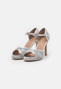 Silberne, glitzernde High-Heelsandalen mit offenem Zehenbereich und Knöchelriemen. Der Absatz ist schmal, und das Obermaterial hat eine schimmernde Textur.