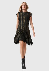 Donna che cammina indossando un vestito di pizzo nero con uno strato sottostante di metallo dorato e stivaletti neri fino alla caviglia su uno sfondo grigio uniforme.