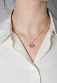 Collana in oro con pendente a forma di farfalla colorata con ali dettagliate, indossata sopra una camicia bianca con colletto.