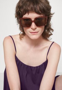 Óculos de sol grandes e quadrados com lentes castanhas escuras e armação bordeaux. A modelo usa uma blusa roxa sem mangas, com cabelo castanho solto e encaracolado.