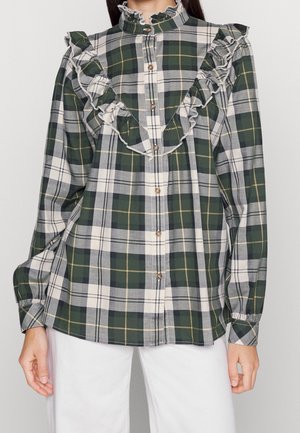 Chemise à carreaux verte, blanche et grise avec volants au niveau de la poitrine et des épaules, portée avec un pantalon blanc.