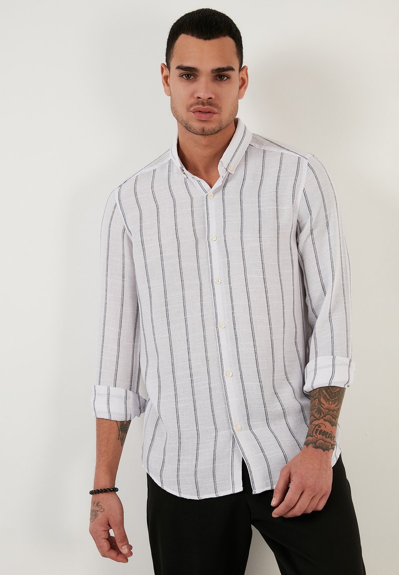 Camicia bianca con righe blu navy, maniche lunghe arrotolate ai gomiti, tessuto leggero e colletto classico.