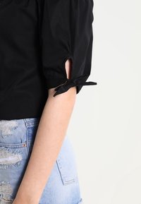 Blusa nera di cotone con maniche a sbuffo, caratterizzata da dettagli a taglio e fiocchi ai polsini. Indossata con pantaloni corti in denim strappati.