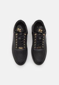 Ett par svarta låga sneakers med gyllene ögla, snören och "FP"-logotyp på den vadderade tungan, visade uppifrån på vit bakgrund.