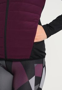 Mano che riposa nella tasca di una giacca a zip color bordeaux e nera, indossata con leggins a motivo geometrico in viola, nero e grigio.
