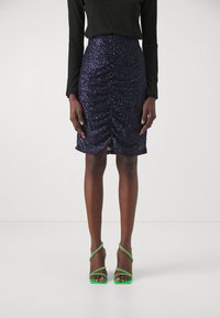 Jupe crayon bleu marine ornée de sequins avec fronces, associée à des sandales à talons hauts vertes à lanières. Silhouette ajustée, détail de surface texturée.