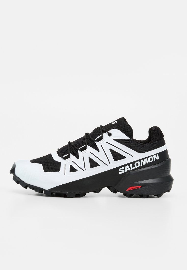 Chaussure de trail running Salomon noire et blanche avec semelle robuste et nom de la marque sur le côté, conçue pour les activités en plein air et l’adhérence.