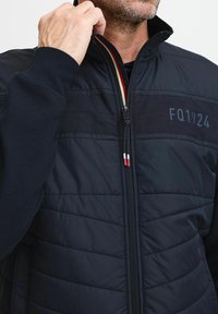 Marineblauer, gesteppten Blouson mit hohem Kragen, Reißverschluss, gerippten Ärmeln und einem aufgedruckten Logo "FQ1924" auf der Vorderseite. Akzente in Rot und Creme.