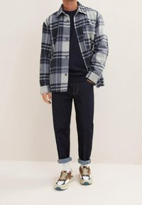 Man som bär en svart och grå rutmönstrad skjorta med knappar över en svart tröja, mörka uppvikta jeans, vita strumpor och beige sneakers med blå detaljer.