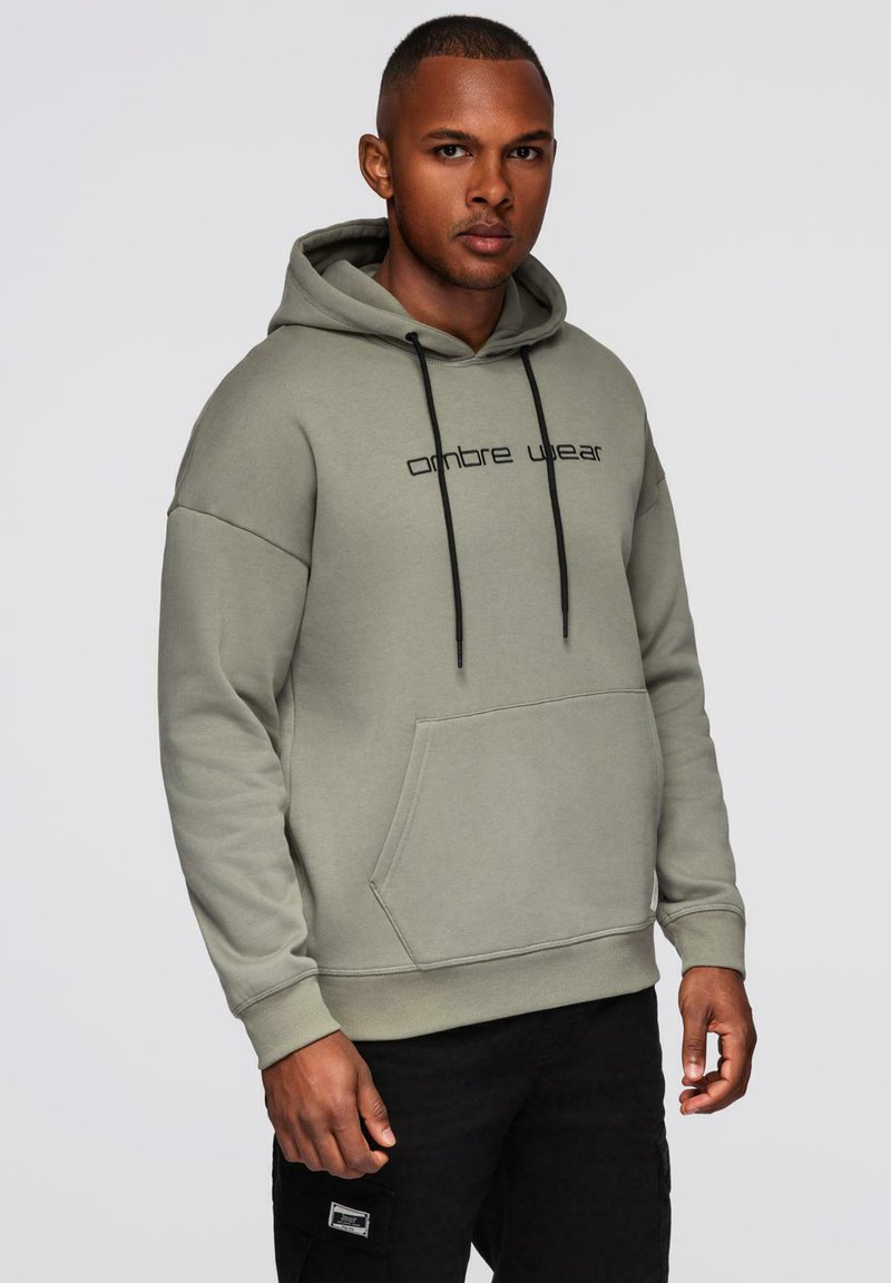 Homme portant un sweatshirt à capuche gris clair avec des cordons de serrage noirs et un texte noir, associé à un pantalon noir, se tenant devant un fond uni.