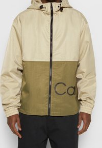 Kurtka typu wiatrówka w beżowym i oliwkowym kolorze, z zamkiem z przodu, regulowanym kapturem oraz logo na dolnym przednim panelu.