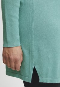 Un top lavorato a maglia verde menta con maniche lunghe, uno spacco laterale sull'orlo e una texture morbida. Il design è semplice e aderente.
