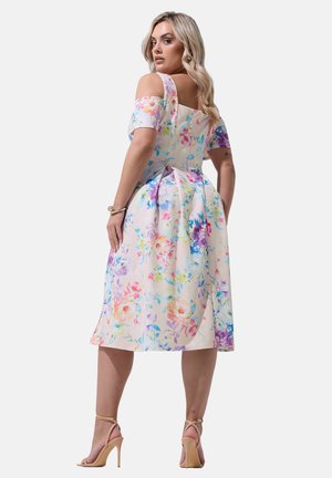 Frau in einem knielangen, schulterfreien Blumenkleid mit Pastellfarben, die hautfarbene High-Heel-Sandalen trägt und mit dem Rücken zur Kamera posiert.