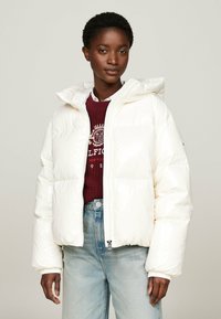 White puffer jacket with a hood, featuring a zip front, ribbed cuffs, and smooth, shiny fabric. Worn over a burgundy knit sweater and denim.