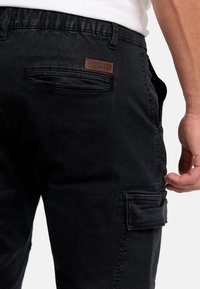 Gros plan sur une personne portant un pantalon cargo noir avec une poche passepoilée à l'arrière et une étiquette de marque rectangulaire marron près de la ceinture.
