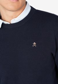 Granatowy sweter z dzianiny o teksturowanym wzorze, z małym haftowanym różą i skrzyżowanymi patykami w jasnym różu na lewej piersi.
