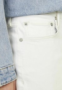 Jeans in denim bianco con design a cinque tasche, con hardware in metallo e dettagli di cucitura a contrasto, abbinati a una camicia in denim azzurro chiaro.