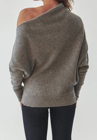 Grauer, übergroßer Strickpullover mit schulterfreiem Design, geripptem Saum und weicher Textur. Trägt ihn mit dunklen Jeans, die eine entspannte Passform zeigen.