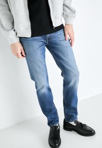 Faded blue denim jeans with a straight fit, worn with a black shirt and grey zip-up jacket, paired with black loafers featuring tassels.