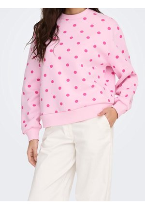 Vrouw draagt een lichtroze sweatshirt met donkerroze stippen en witte broek, één hand in de zak, voor een effen achtergrond.