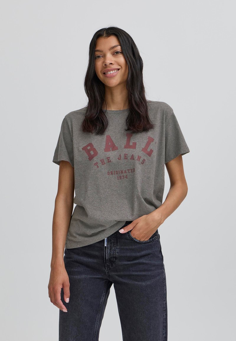 Grå bomulds t-shirt med korte ærmer, der har rød tekst: "BALL THE JEANS" og "ORIGINATED 1974." Bæres med mørke jeans. Casual pasform.