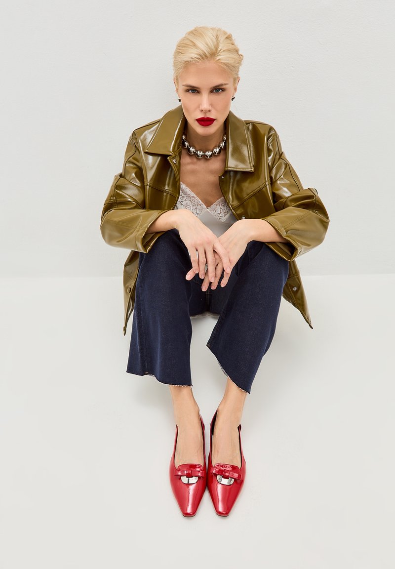 Giacca in pelle verniciata verde oliva, top in pizzo bianco, pantaloni cropped in denim scuro, e scarpe rosse lucide con punta a punta e accento metallico.
