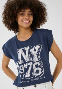 Tumši zila īsām piedurknēm t-krekls, izgatavots no kokvilnas, ar baltu grafisku apdruku, kurā ir teksti "NY 1976" un "Nacionālā čempionāta".