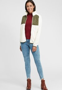 Hvit fleecejakke med grønne quiltede skulderpaneler, glidelås foran, kombinert med en burgunder høyhalset genser, lyseblå jeans og brune ankelstøvler.